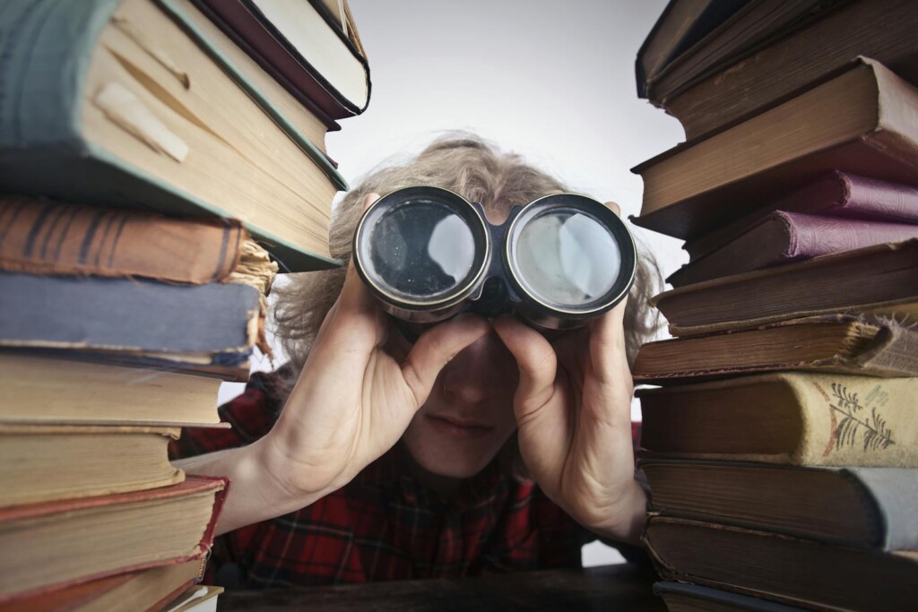 A man observing through binoculars surrounded by stacks of vintage books indoors.